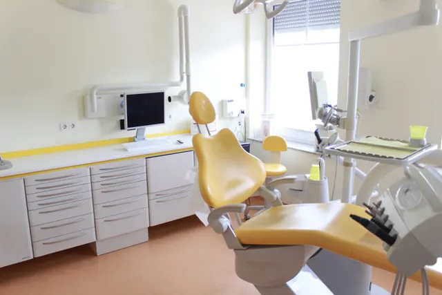 Behandlungszimmer der Praxis mit gelbem Behandlungsstuhl, weißen Schränken und Computerarbeitsplatz in hellem Licht