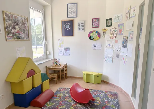 Kinderecke im Wartezimmer mit Spielteppich, roter Wippe, Kindertisch und selbstgemalten Bildern unter einem großen Fenster